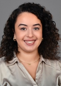 csm_sen-melisa-6946-2_773fec1fe8 Lächelnde Frau mit lockigem dunklem Haar, die eine beigefarbene Bluse, eine goldene Halskette und Reifohrringe trägt, auf grauem Hintergrund.